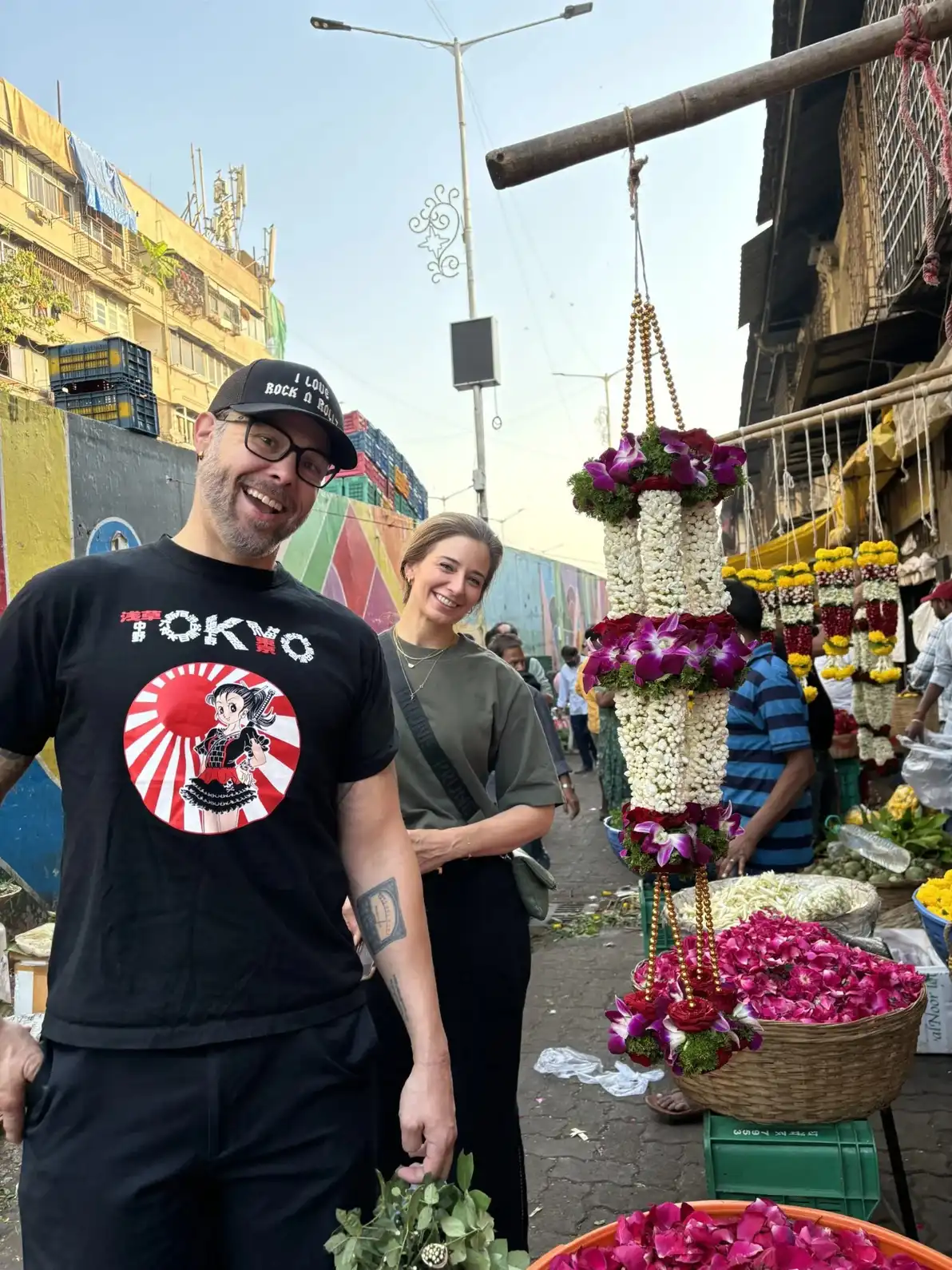 Smiling travellers at Mumbai's flower market with hanging jasmine and rose garlands on display ; Cityscape mumbai tours.