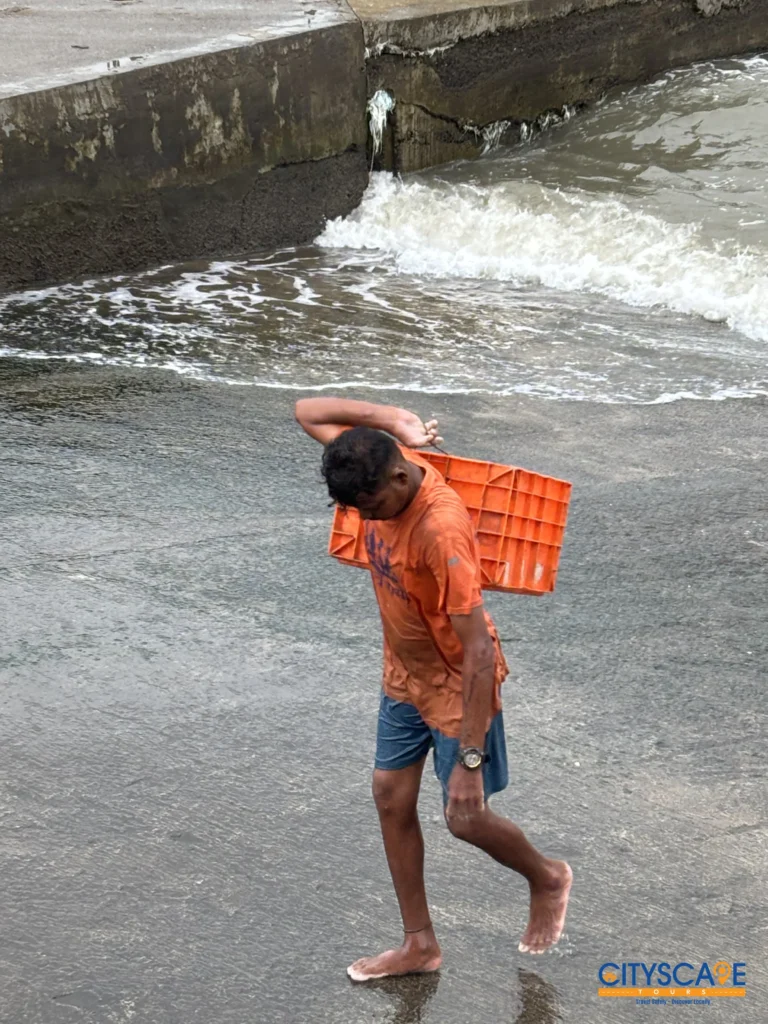 Barefoot fish market worker in an orange T-shirt carrying a plastic crate on his back along a wet slipway as waves wash in, on the Mumbai dawn tour by Cityscape Mumbai Tours.
