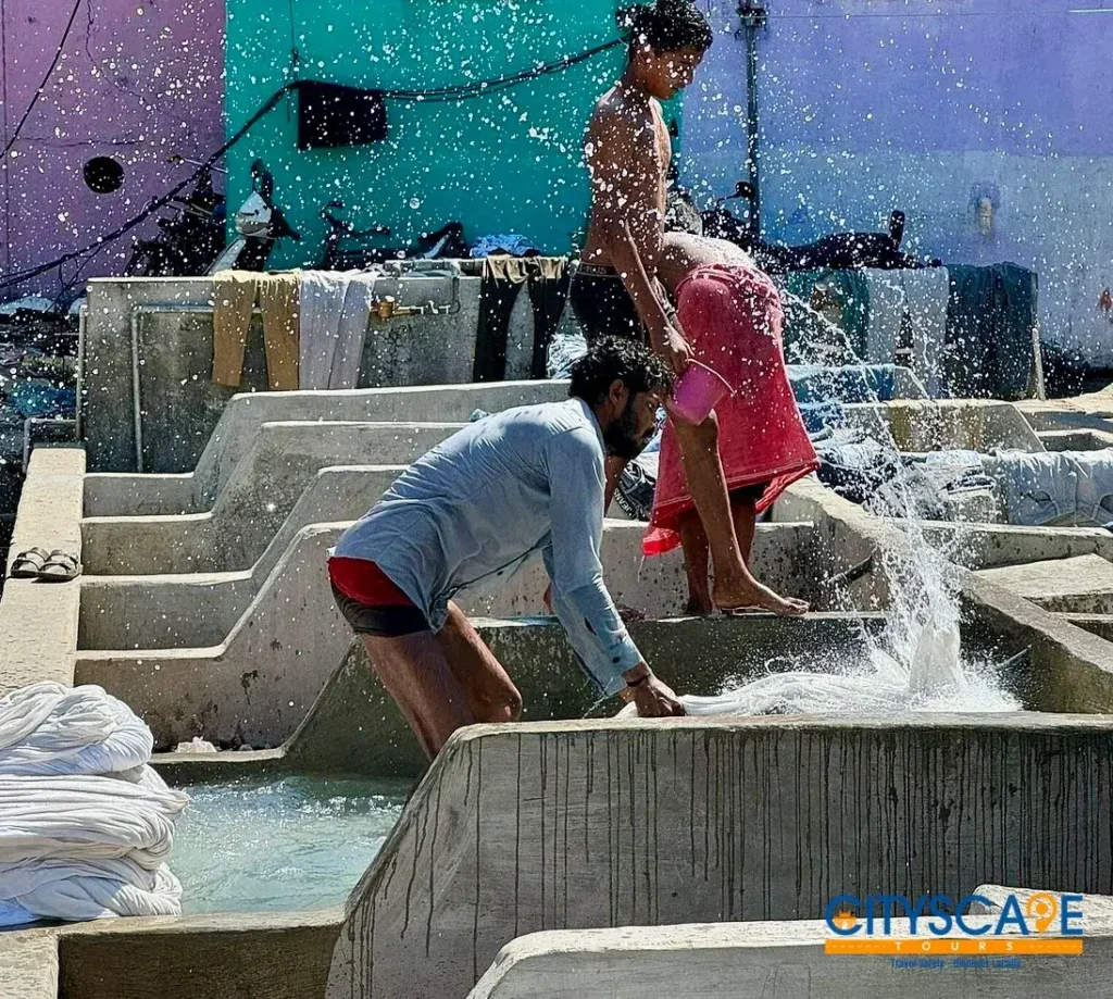 Dhobi Ghat workers washing clothes by hand in concrete wash pens with water splashing, in front of colourful houses, on the Mumbai dawn tour by Cityscape Mumbai Tours.