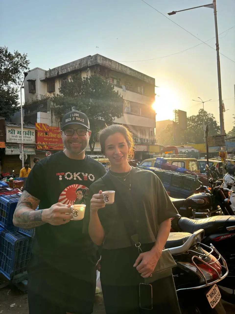 Two smiling guests holding small cups on a busy market street in the warm early morning sun, on the Mumbai dawn tour by Cityscape Mumbai Tours.