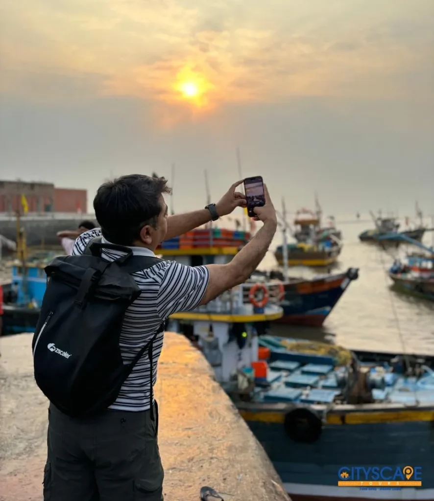 Visitor taking a phone photo of the golden morning sun over the harbour and fishing boats, on the Mumbai dawn tour by Cityscape Mumbai Tours.