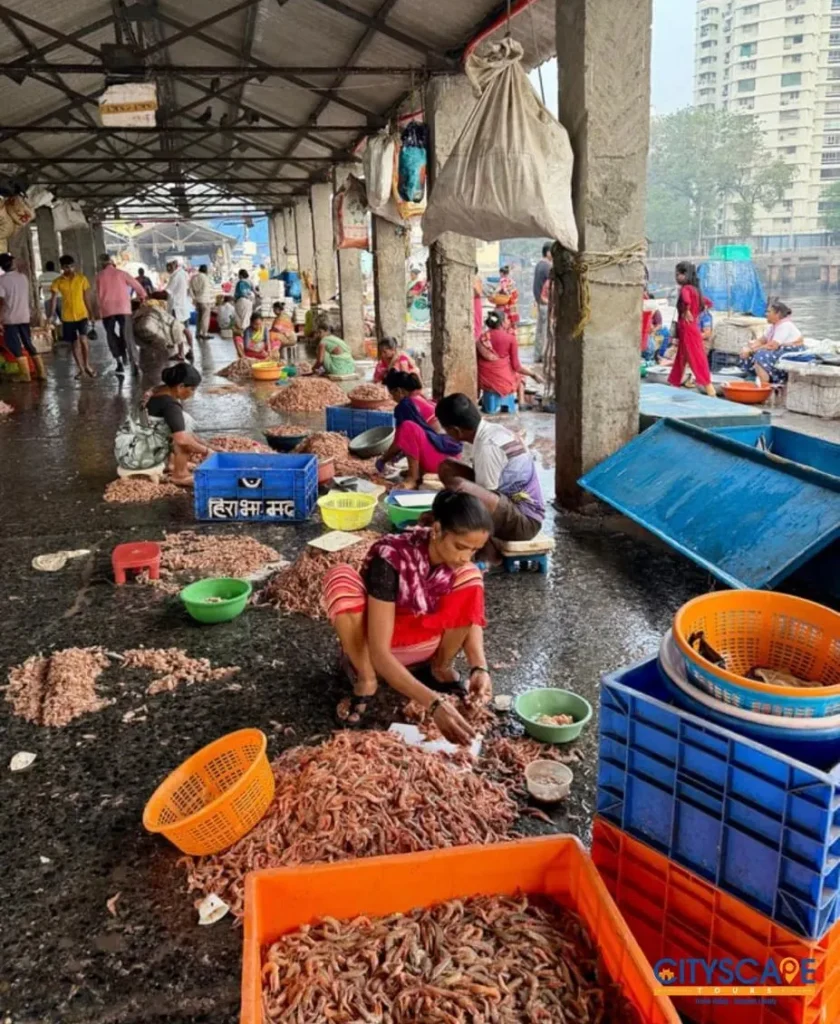 Women sitting on the wet floor of a covered fish market shed, sorting and peeling prawns into baskets and crates, on the Mumbai dawn tour by Cityscape Mumbai Tours.